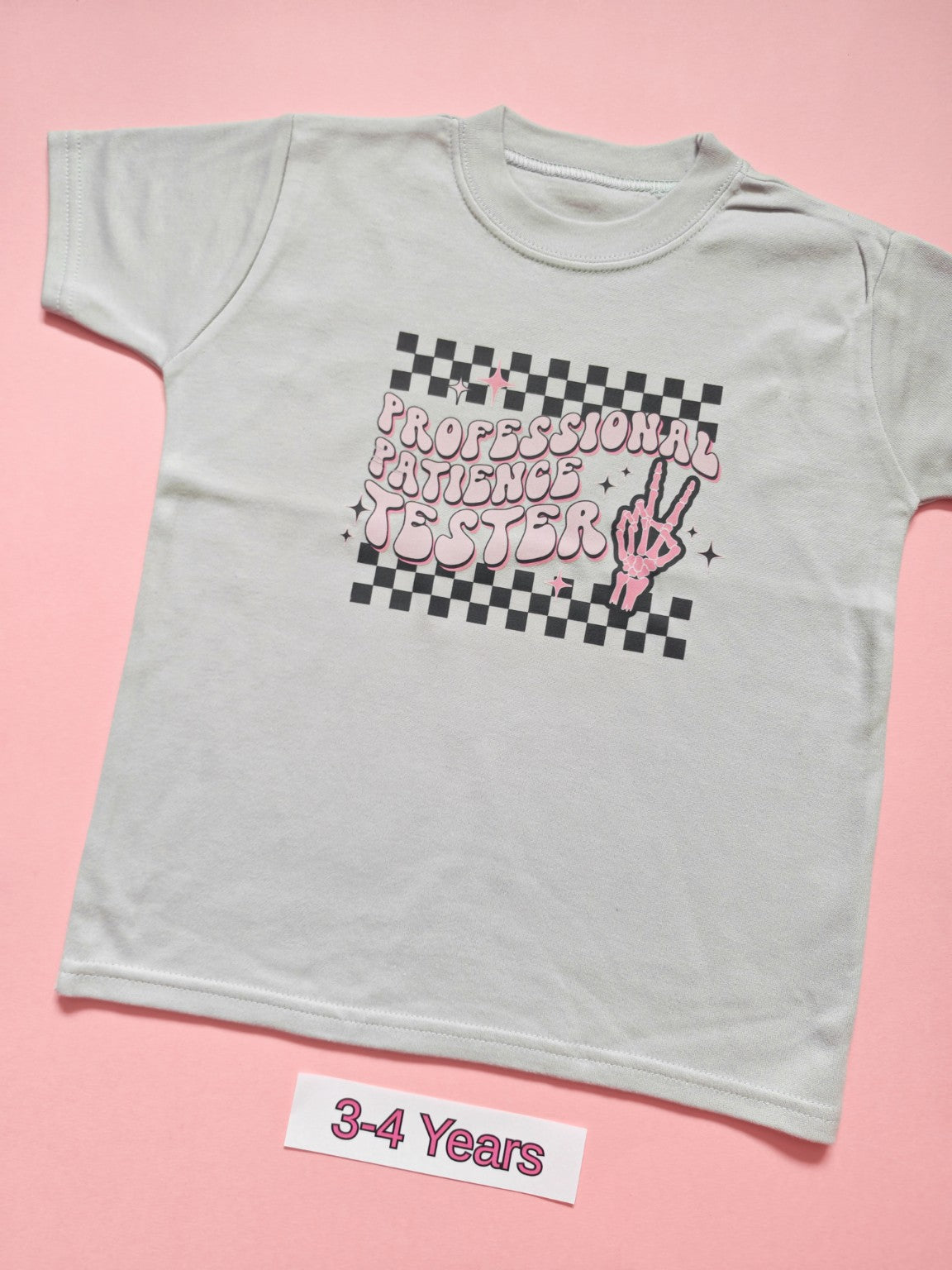 White children's T-shirt with pink Professional Patience Tester checkerboard design, size 3-4 years